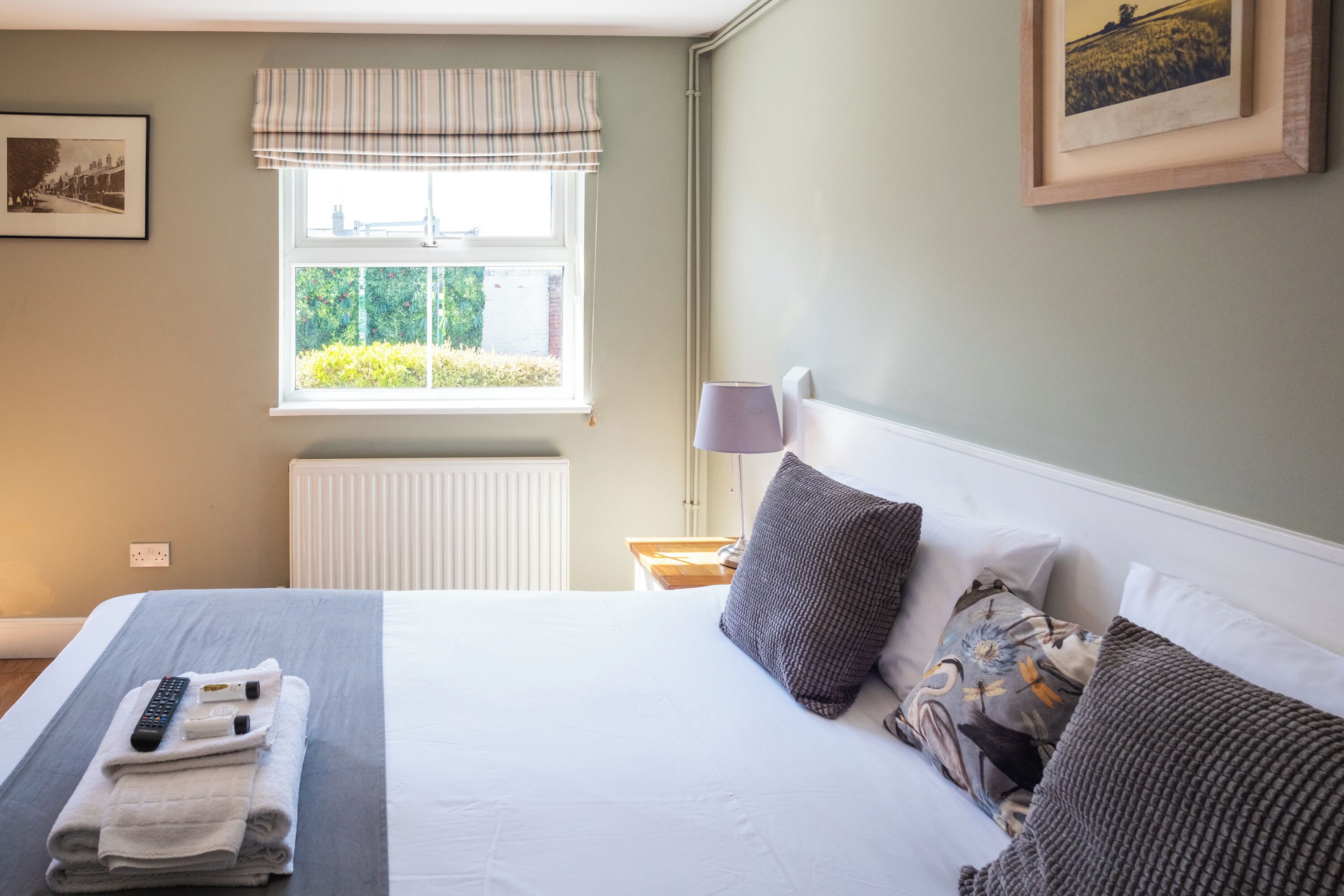 A neatly made bed with a remote control and towels, a window with a view of greenery, and a framed picture on the wall.