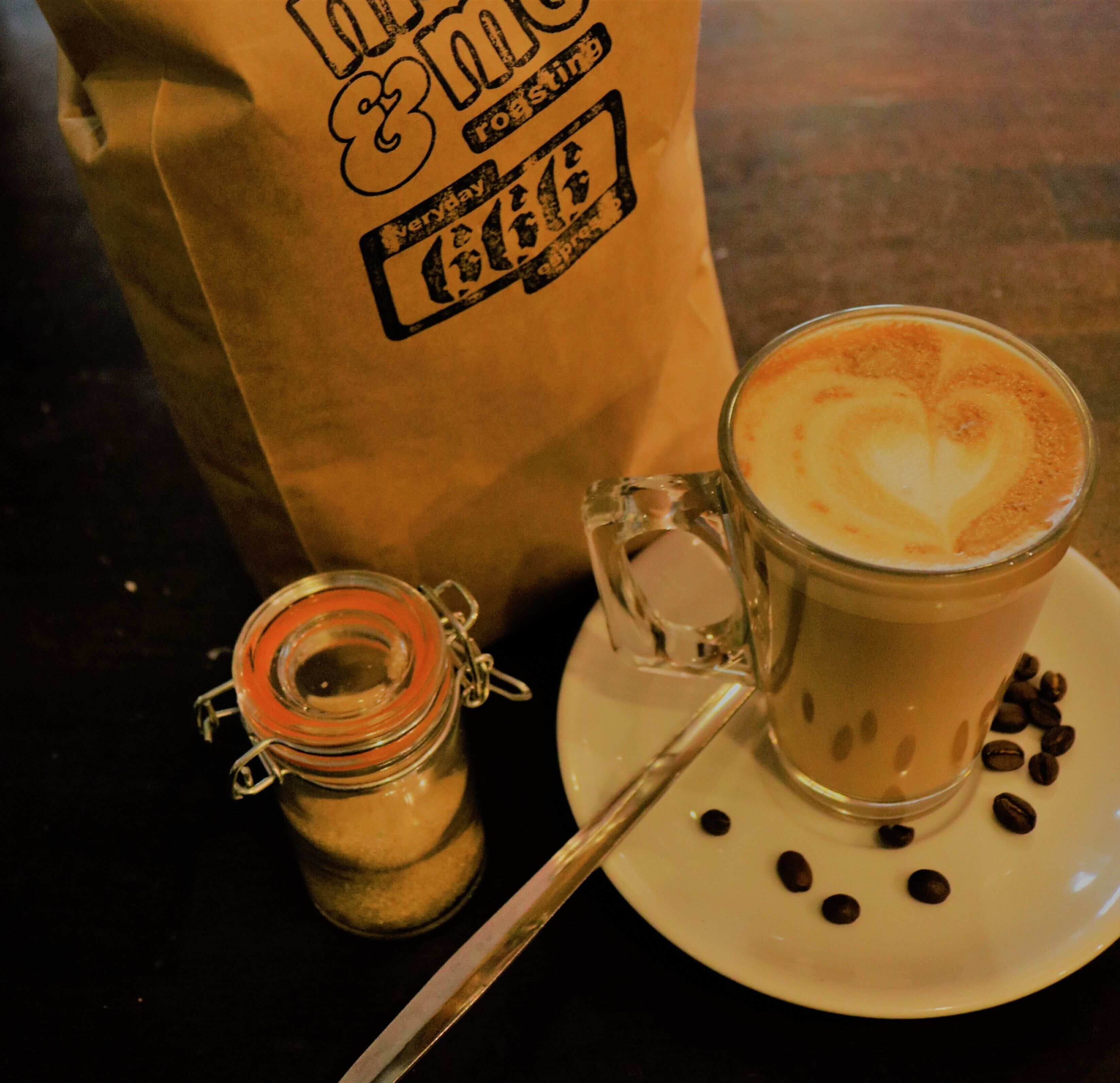 A cup of coffee with a heart design on the foam, a spoon, coffee beans, and a paper bag with text.