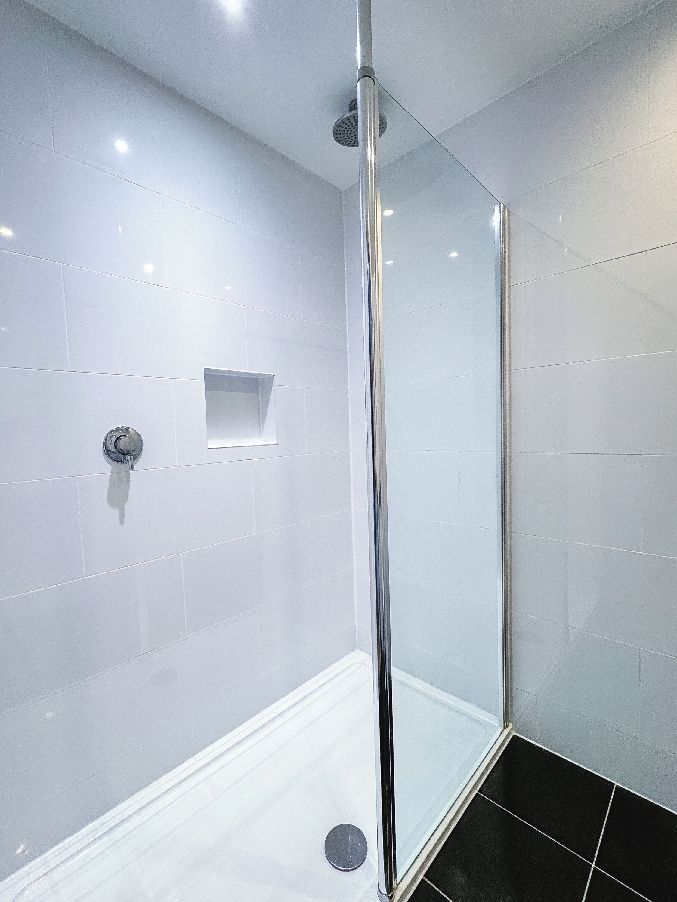 Um chuveiro moderno com porta de vidro, paredes revestidas de azulejos brancos e chão de azulejos pretos.