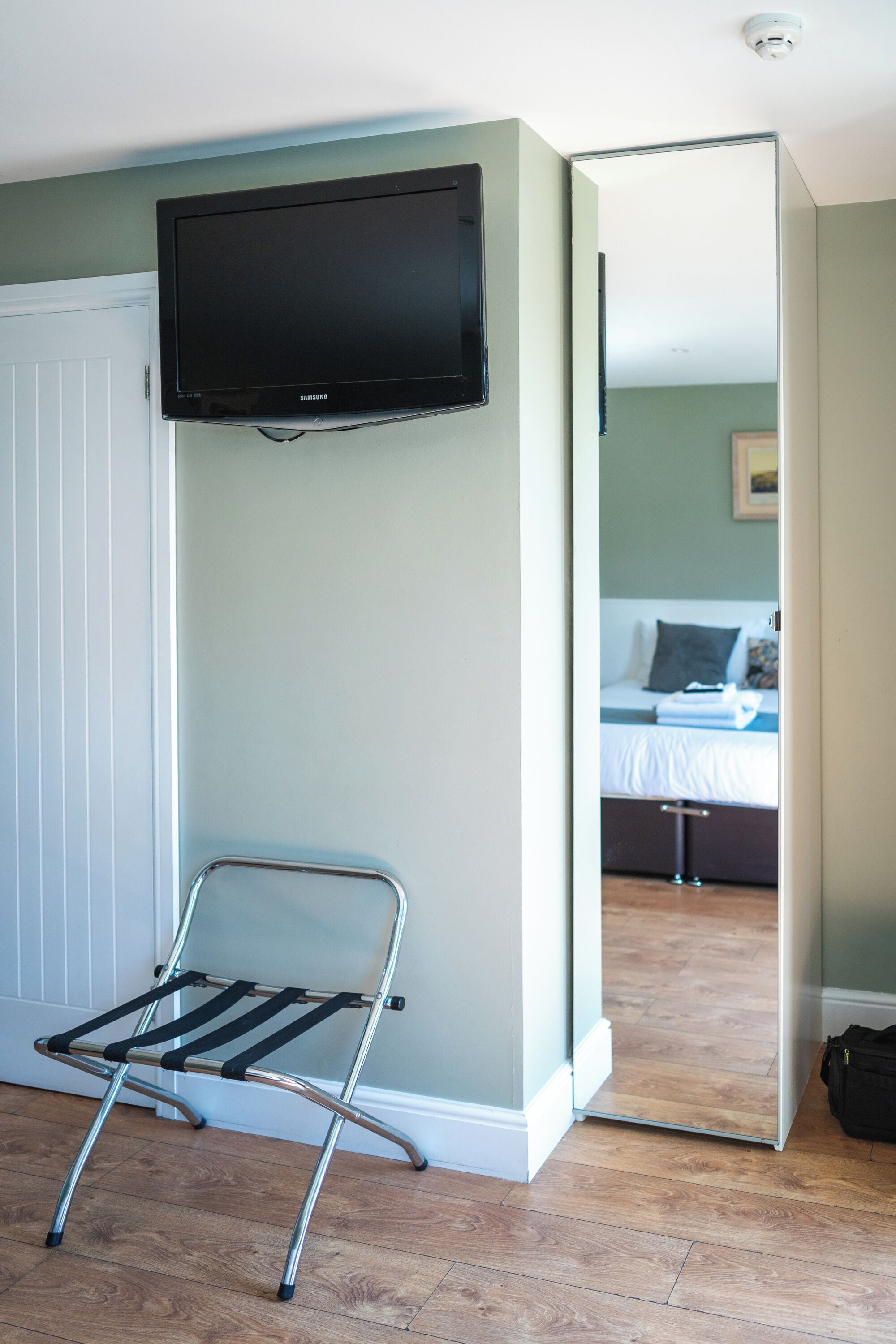 Una stanza con una televisione montata a parete, una sedia pieghevole in metallo e un letto visibile attraverso una porta aperta.