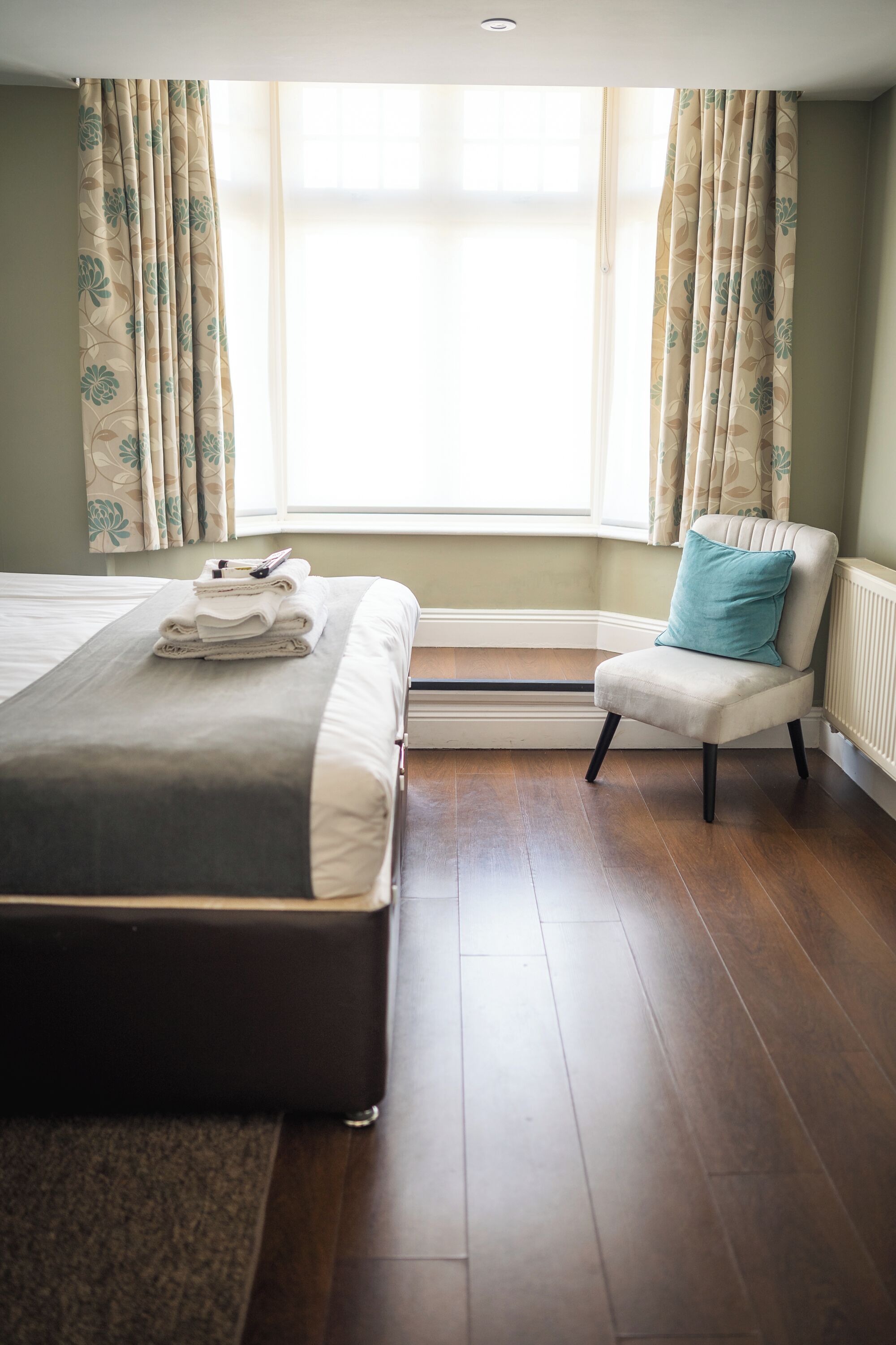A neatly made bed with a folded towel and a pillow, a chair with a cushion, and a radiator under a window with curtains.