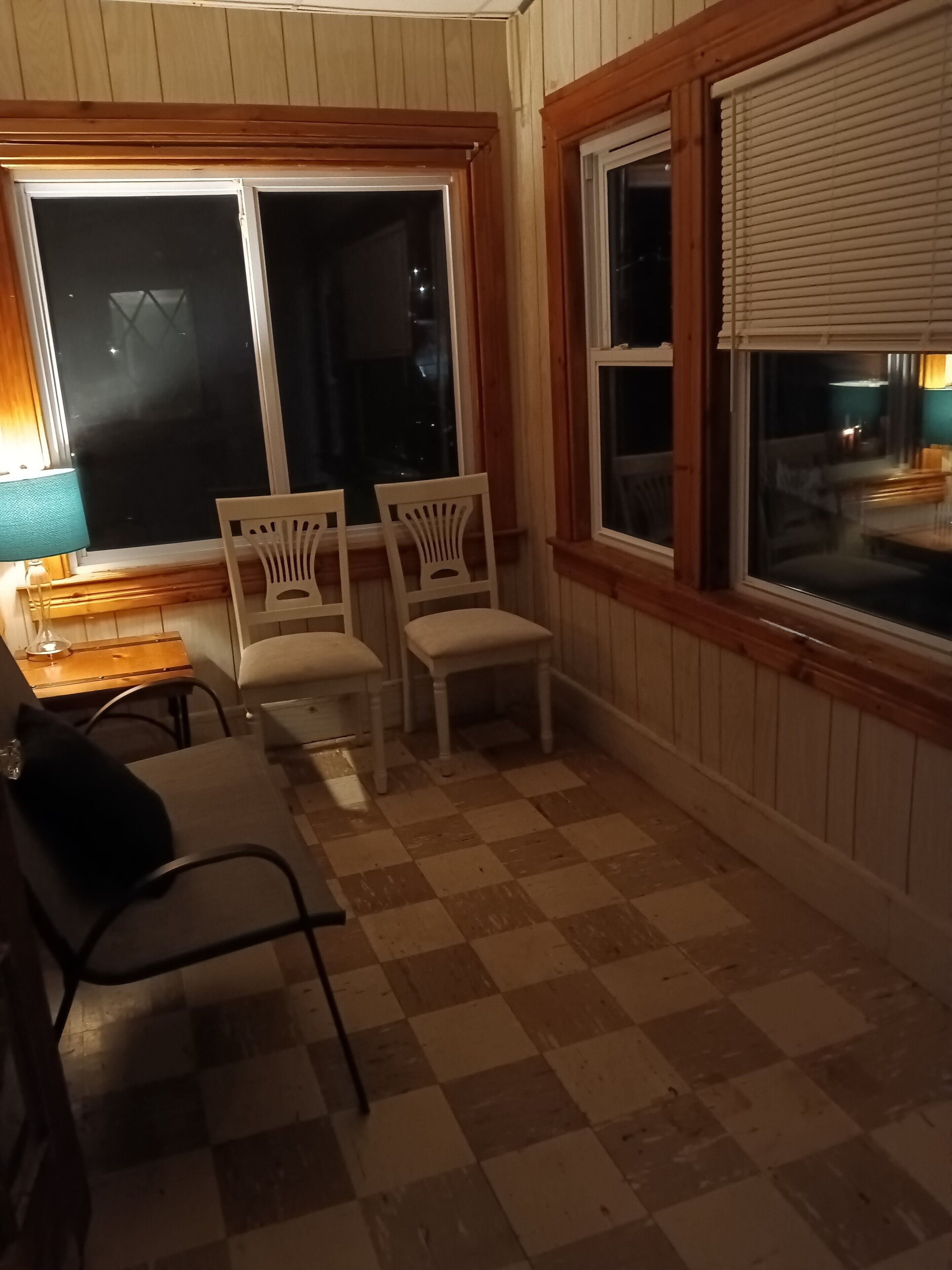 Een gezellige kamer met een zwart-wit geruit vloerkleed, houten wanden en een raam met luiken. Er staan twee witte stoelen en een tafel met een lamp.