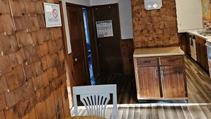 A room with a wooden wall, a dining table, a white chair, a kitchen area with wooden cabinets, and a sign on the wall. - Indian Lake House (Worcester)