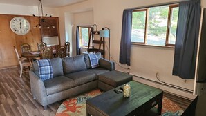 A living room with a sectional sofa, a coffee table, a dining table with chairs, a clock, and a window with curtains. - Indian Lake House (Worcester)