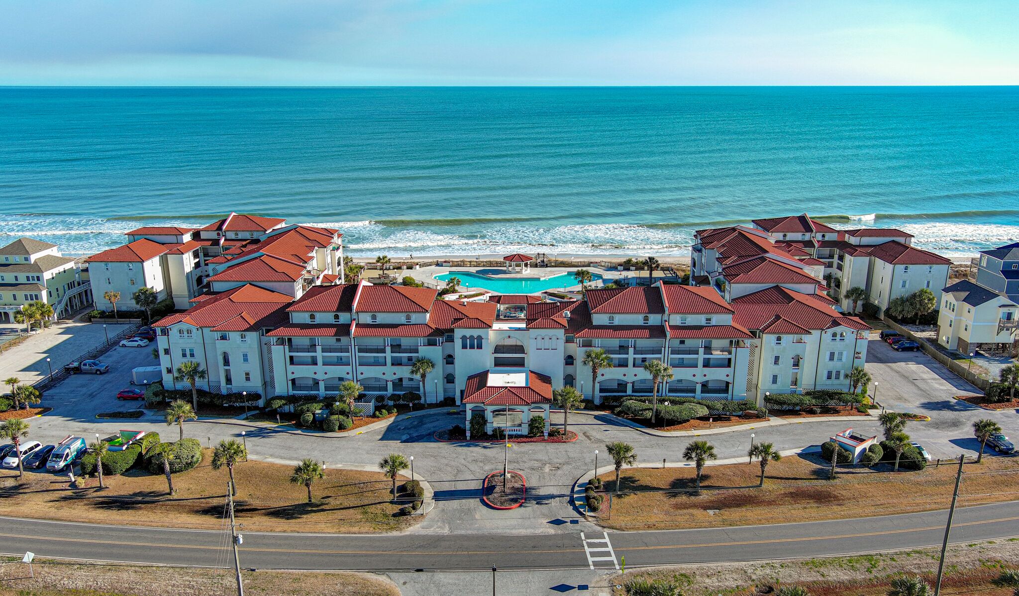 A coastal hotel with multiple buildings, a central swimming pool, and a beach view.
