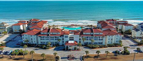 A coastal hotel with multiple buildings, a central swimming pool, and a beach view.
