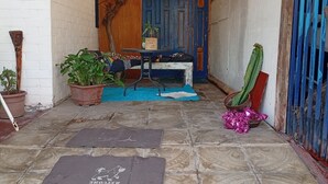 A covered patio area with a wooden ceiling, potted plants, a table with a chair, and a blue door.