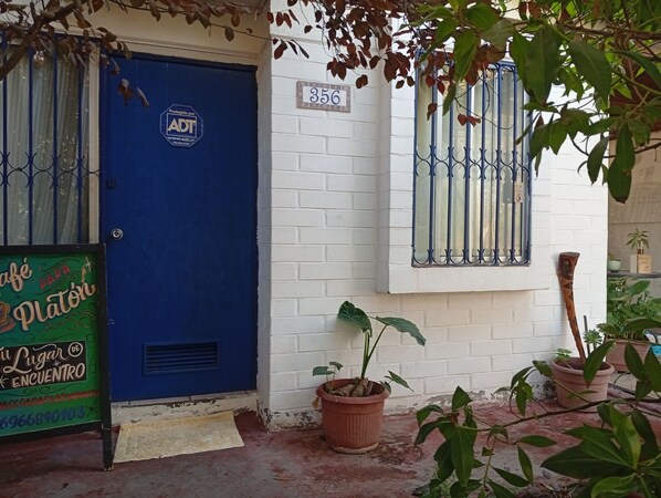 A blue door with a security sign, a white brick wall, a window with metal bars, and potted plants.