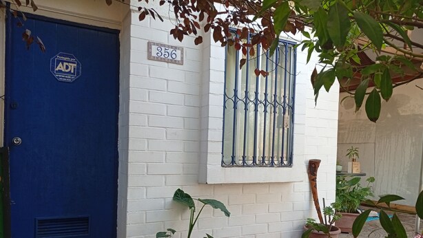 A blue door with a number plate, a white brick wall, a window with metal bars, and potted plants.