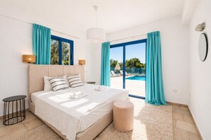 A bedroom with a bed, a bedside table, a lamp, a large window, and a view of a swimming pool. - Villa Estia - Three Bedroom Villa, Sleeps 7 (Peyia)