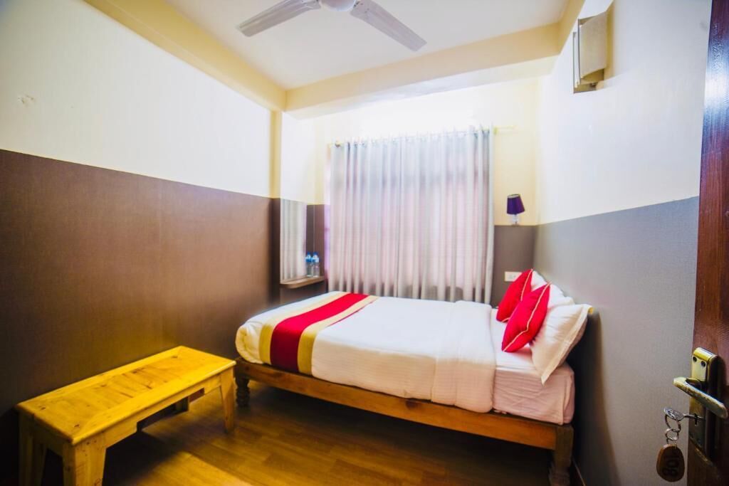 A single bed with a red and white bedspread, a wooden bench, a ceiling fan, and a window with curtains.