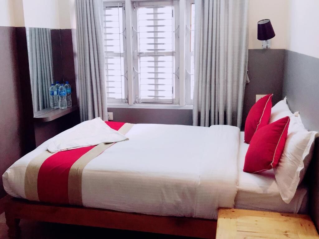 Una cama bien hecha con ropa de cama blanca y roja, un banco de madera, una ventana con cortinas y una lámpara fijada a la pared.