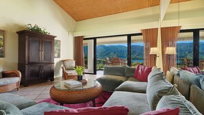 A spacious living room with a large sofa set, a dark wooden cabinet, and a glass coffee table. A large window offers a view of mountains and a lake.