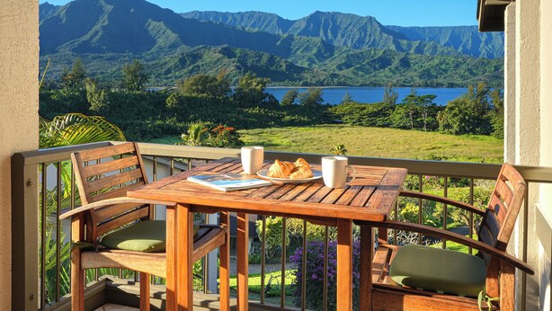 A wooden table with chairs and a plate of croissants, a book, and two cups on a balcony overlooking a mountainous landscape and a lake.