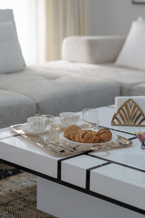 Una bandeja de desayuno con croissants y tazas sobre una mesa de centro blanca en una sala de estar moderna, con un sofá y decoración mural.