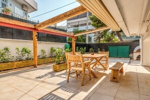 A covered patio with a wooden table and chairs, surrounded by planters and a tiled floor. - Voula Luxury Modern Living with Garden (Vari-Voula-Vouliagmeni)