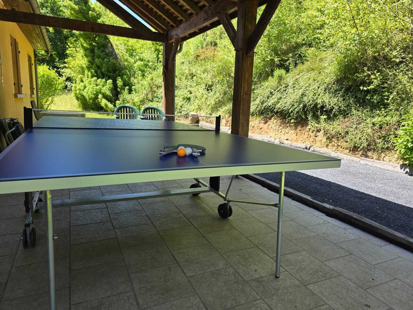 Ein Tischtennis-Tisch im Freien unter einer Holzpergola, mit einem grünen Stuhl und einem Schl äger.