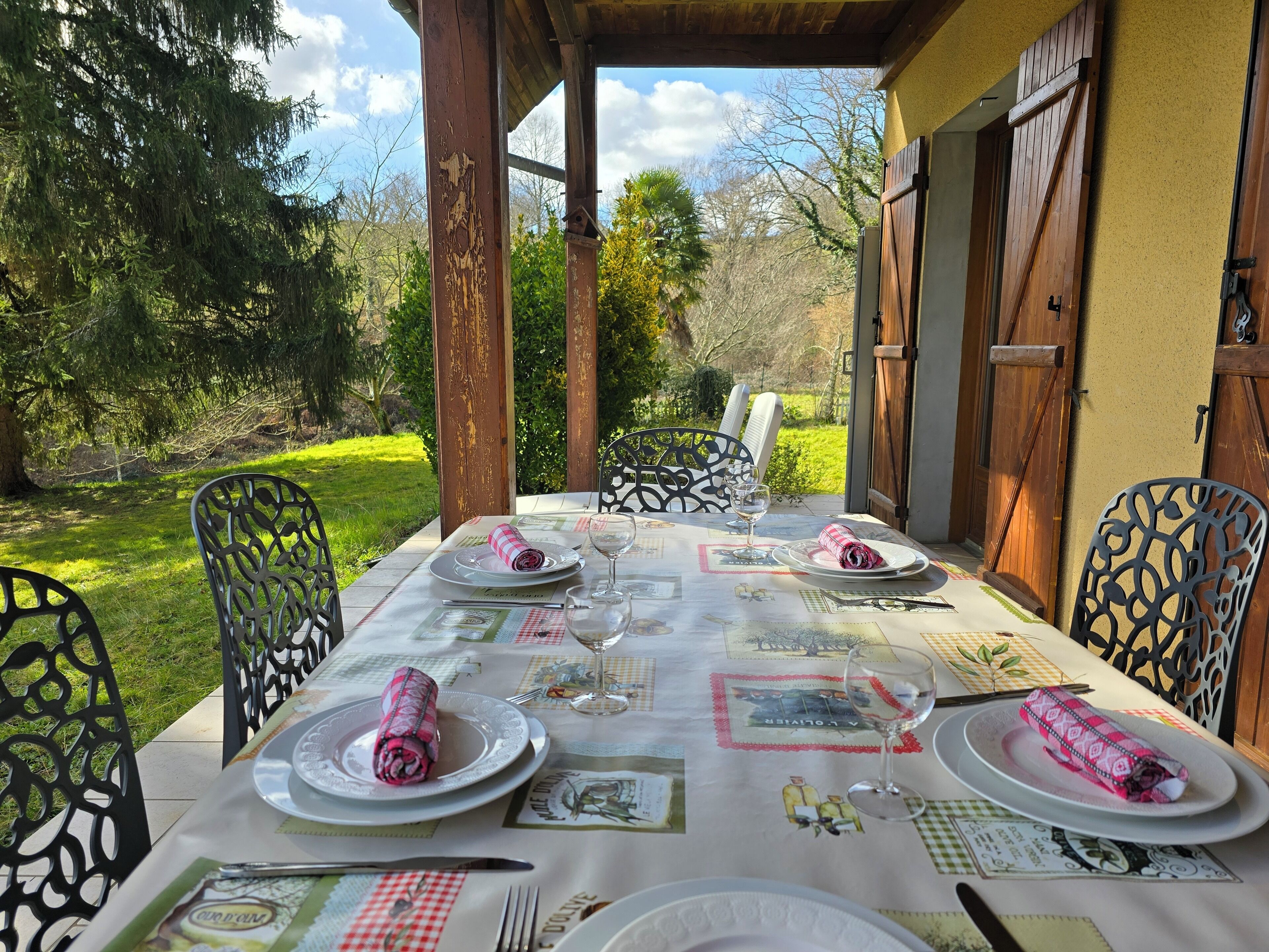 Zona pranzo all'aperto con tavolo imbandito per due, circondata da sedie e vista su un giardino.