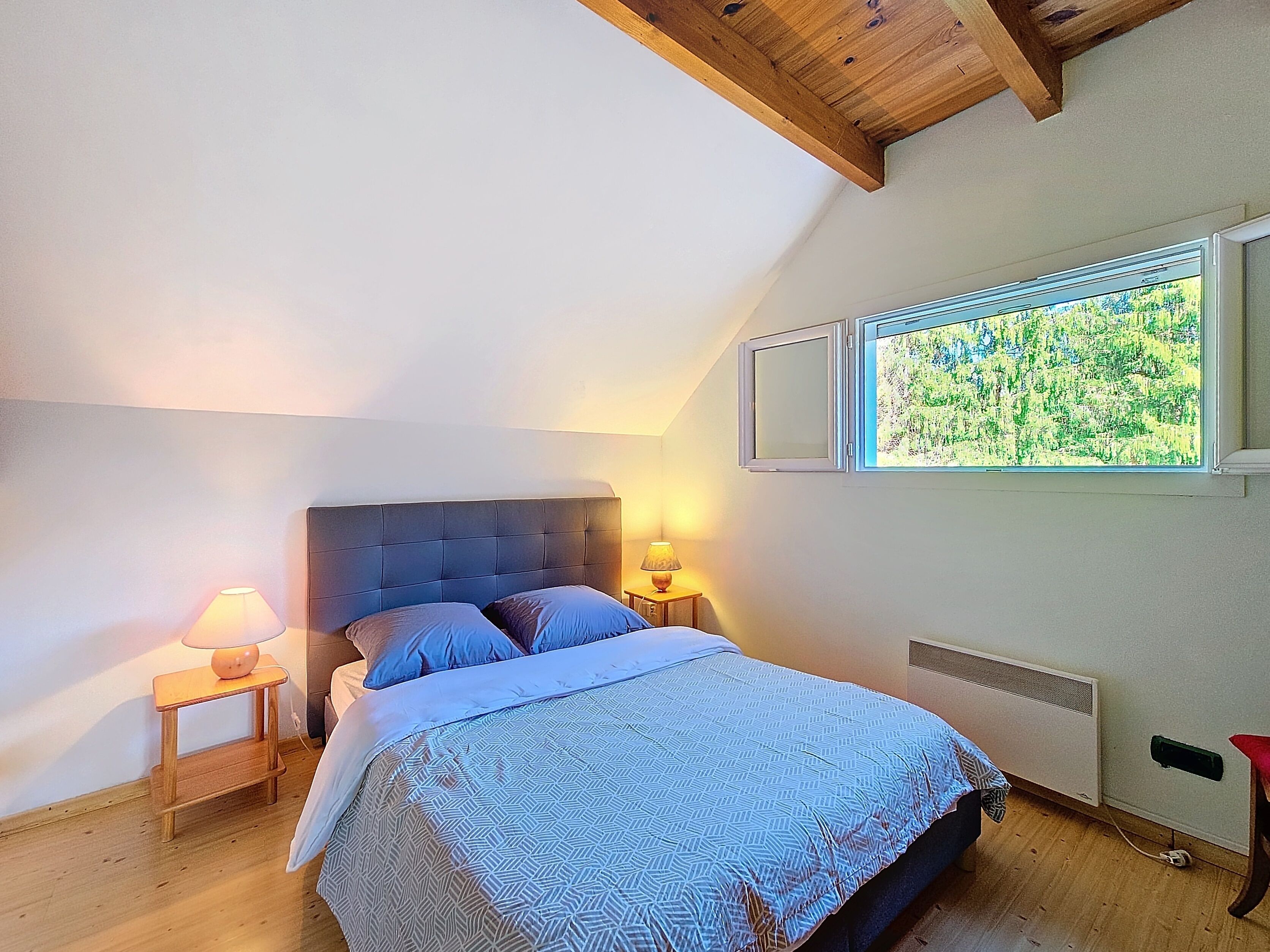 Une chambre avec un plafond en pente, un lit recouvert d’une couvre-lit à motifs, deux lampes de chevet et une fenêtre donnant sur un paysage verdoyant.