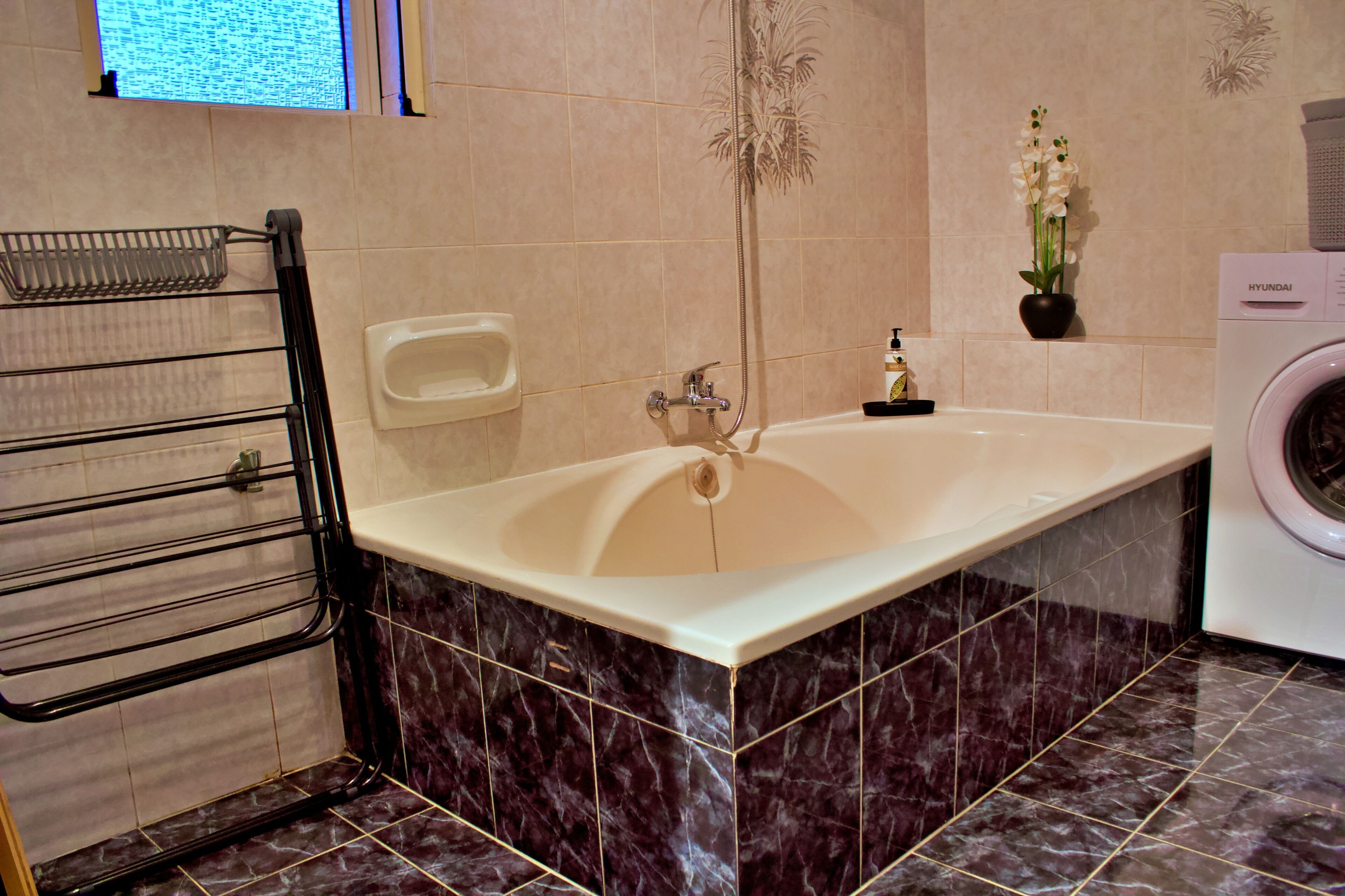 Une salle de bain moderne avec une grande baignoire, un sèche-mains, un lave-linge et une petite étagère ornée de fleurs.