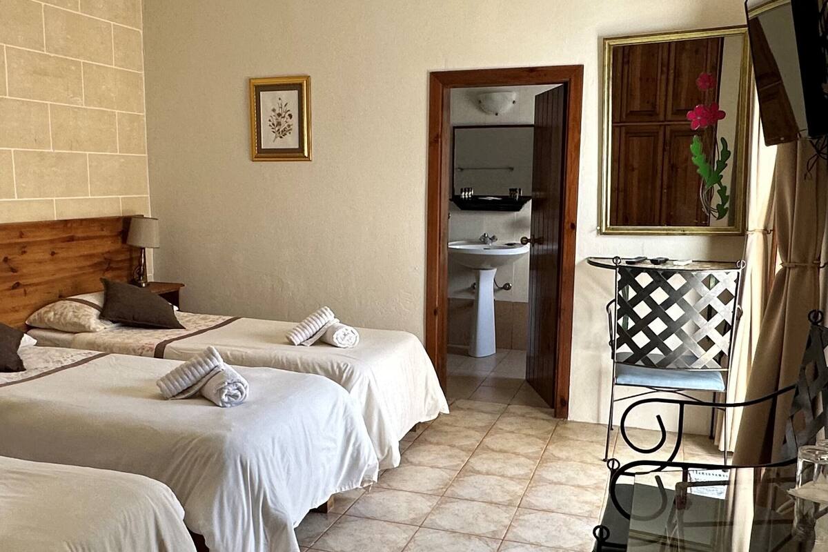 A hotel room with two beds, a wooden headboard, a bedside table with a lamp, a framed picture on the wall, a bathroom with a sink and mirror, and a small table with a decorative item.