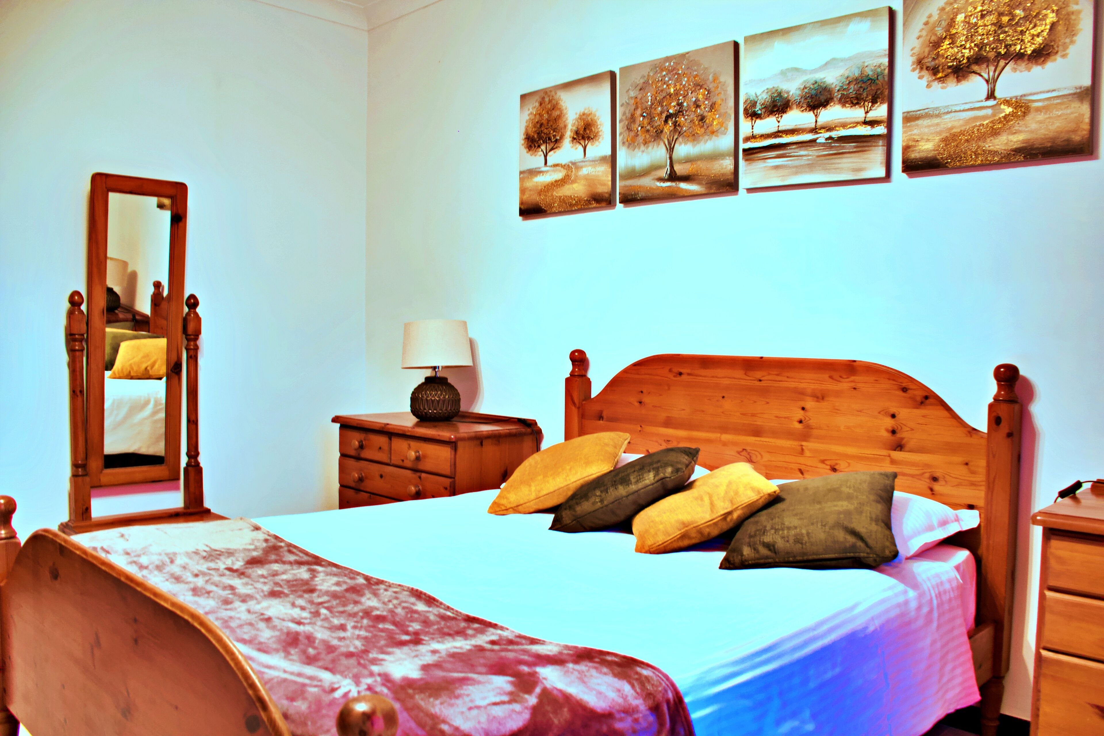 A bedroom with a wooden bed, a nightstand with a lamp, a dresser, and a mirror. There are four framed pictures on the wall.