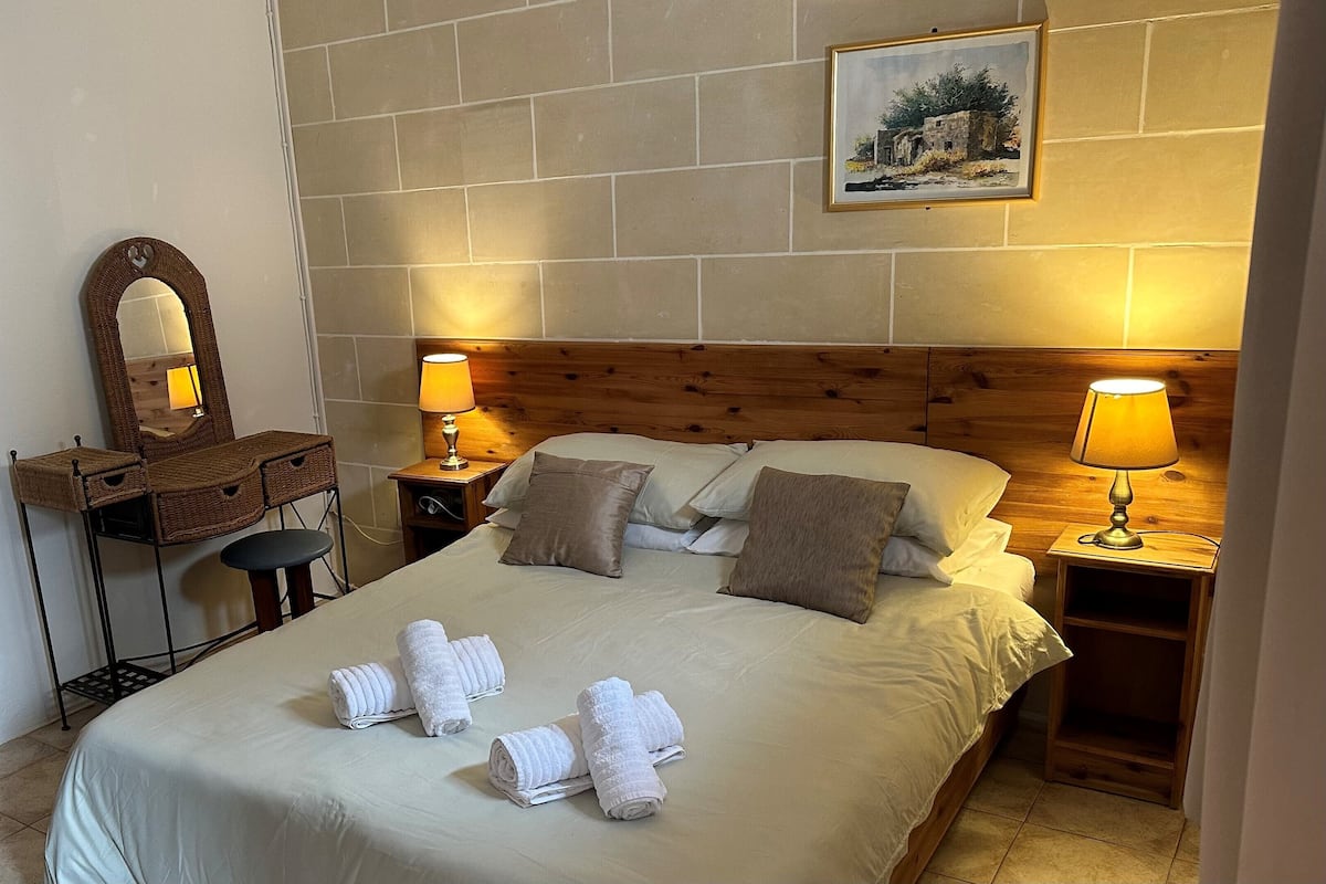 A neatly made bed with towels, a wooden headboard, two bedside lamps, a nightstand, a mirror, and a picture on the wall.