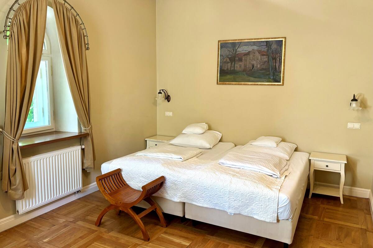 A bedroom with a double bed, two bedside tables, a chair, a radiator, and a painting on the wall.