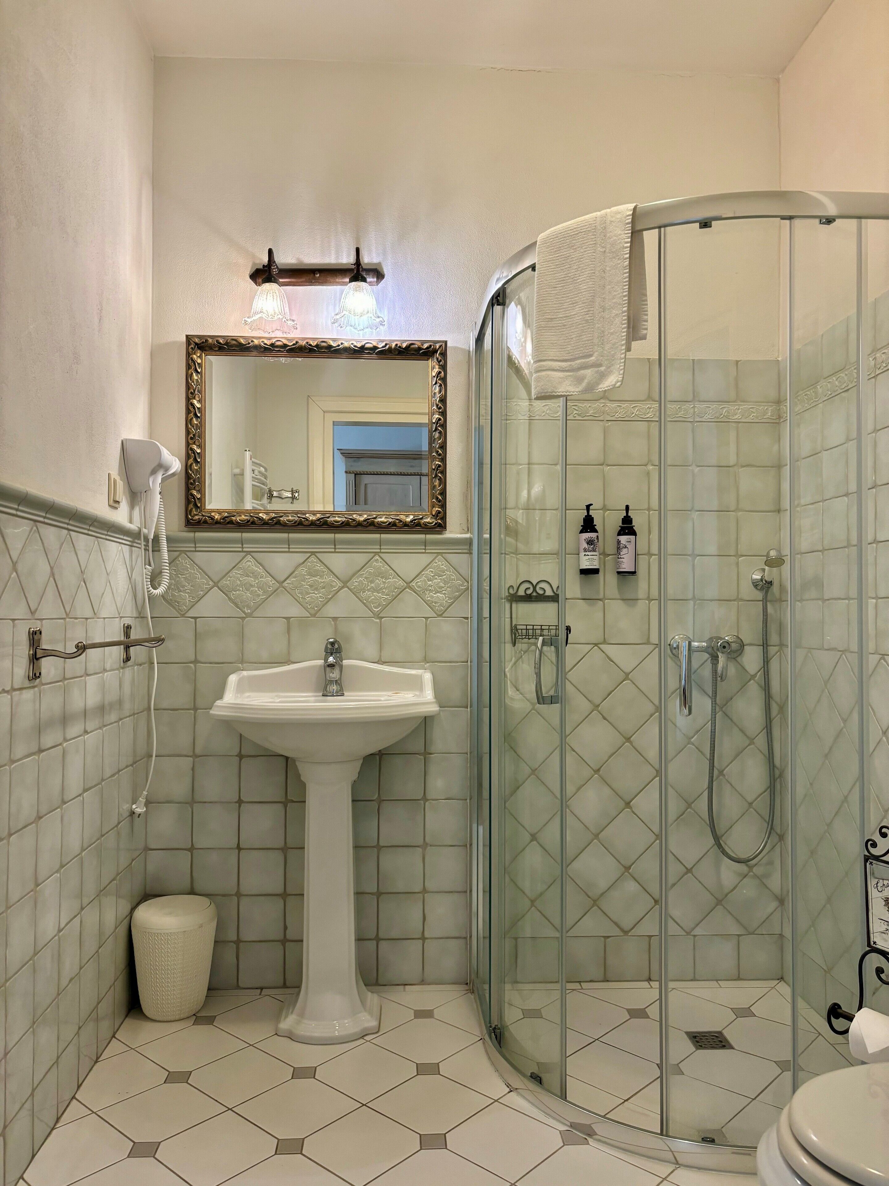 A bathroom with a glass-enclosed shower, a pedestal sink, and a mirror with an ornate frame.