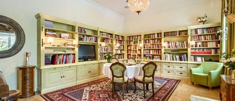 A library with bookshelves, a chandelier, a dining area with chairs, and a green armchair.