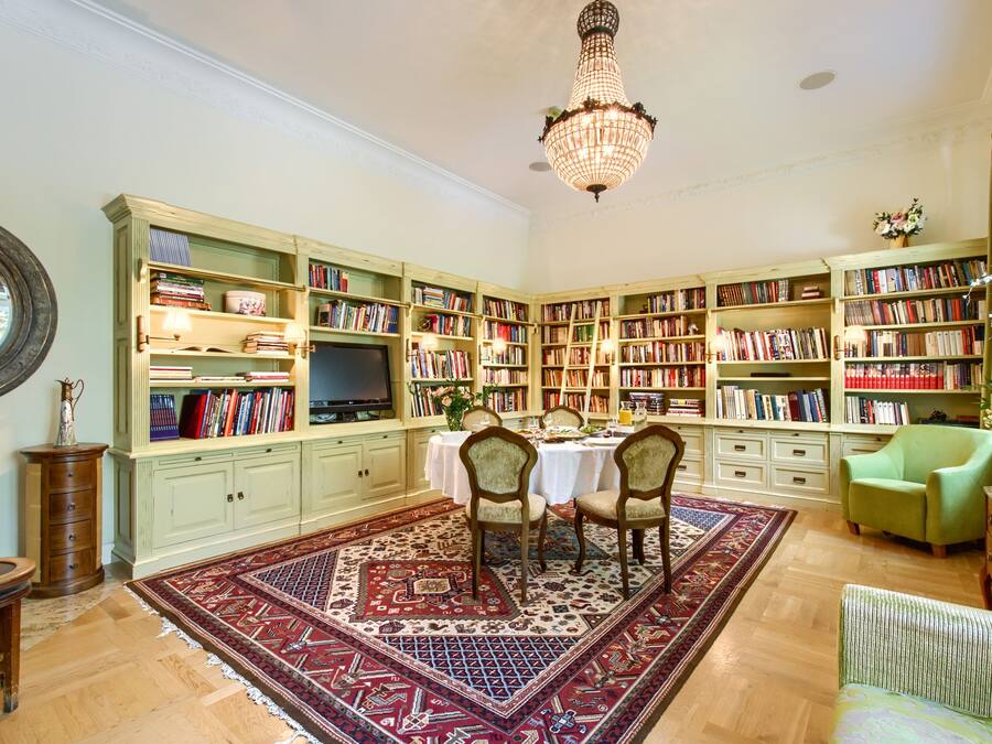 A library with bookshelves, a chandelier, a dining area with chairs, and a green armchair.