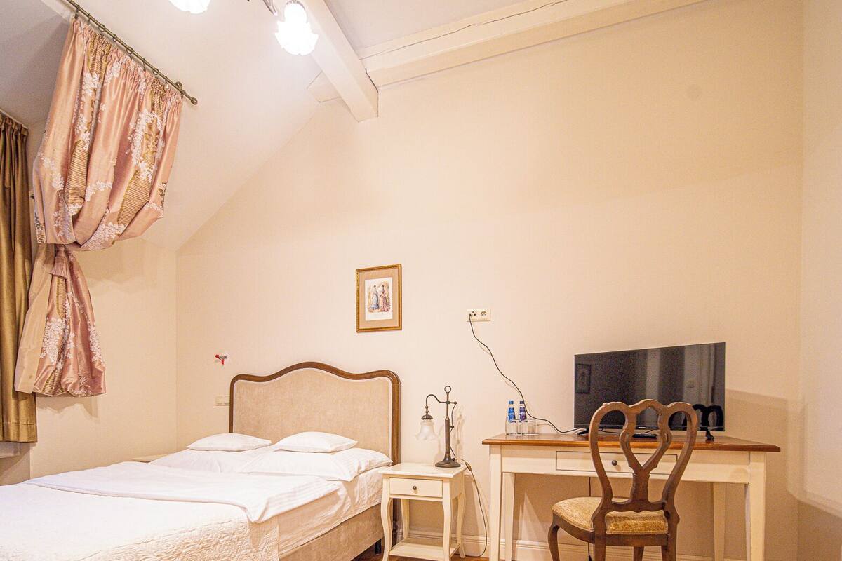 A bedroom with a bed, a desk with a chair, a television, and a chandelier.