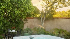 A hot tub in a backyard with a stone patio, surrounded by greenery and a wall.