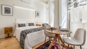 Un dormitorio con cama, mesitas de noche, una mesa redonda con sillas y un ventanal grande.