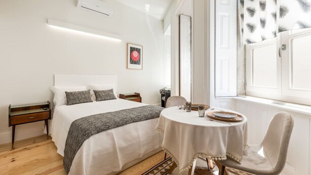 Une chambre à coucher comprenant un lit, une table de chevet, une chaise, une table ronde recouverte d’une nappe blanche et une fenêtre agrémentée de rideaux à motifs.