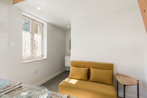 Um quarto com sofá, uma mesa pequena, janela com cortinas e parede branca.