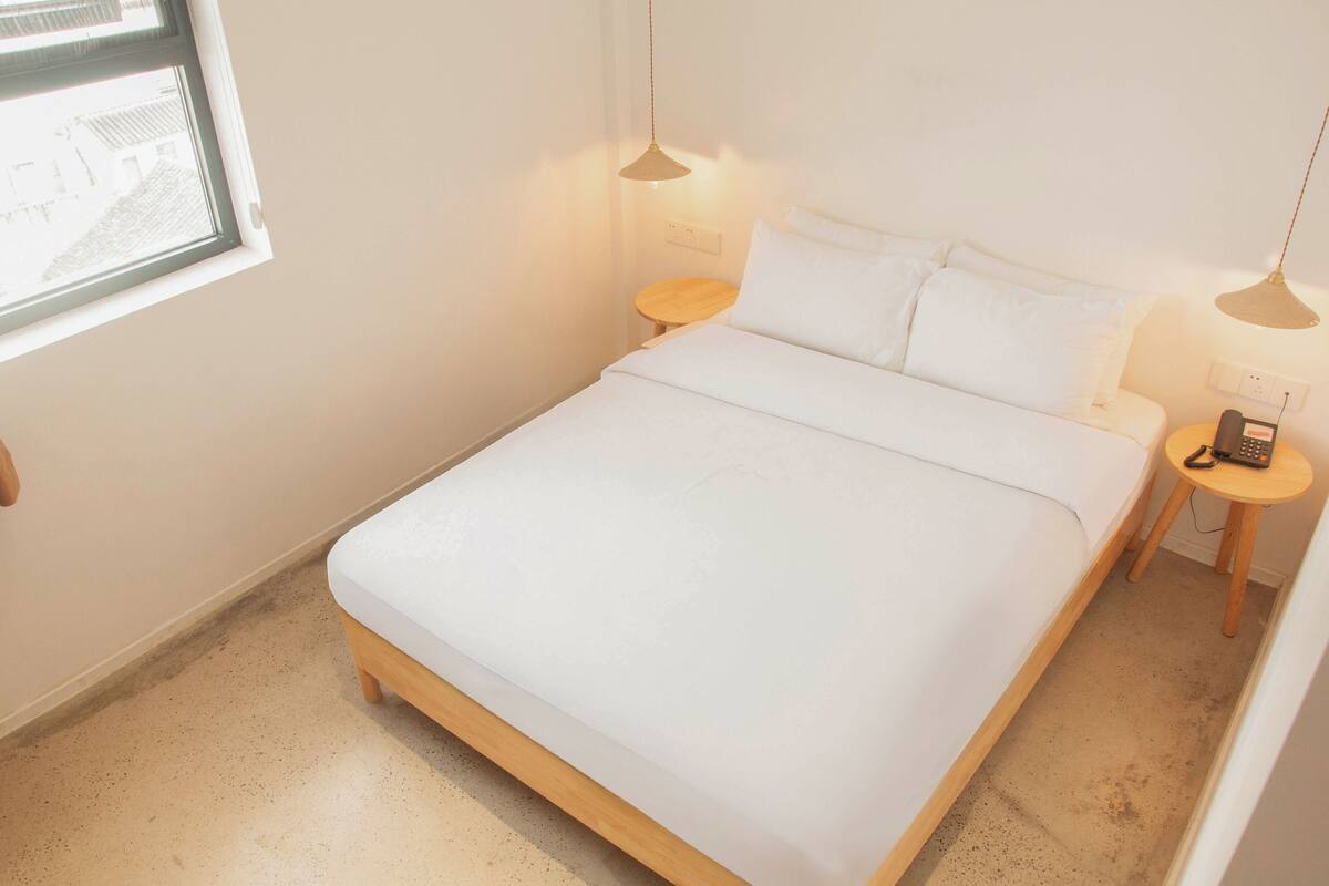 A neatly made bed with white linens, a small wooden bedside table with a lamp, and a window with a view of buildings.