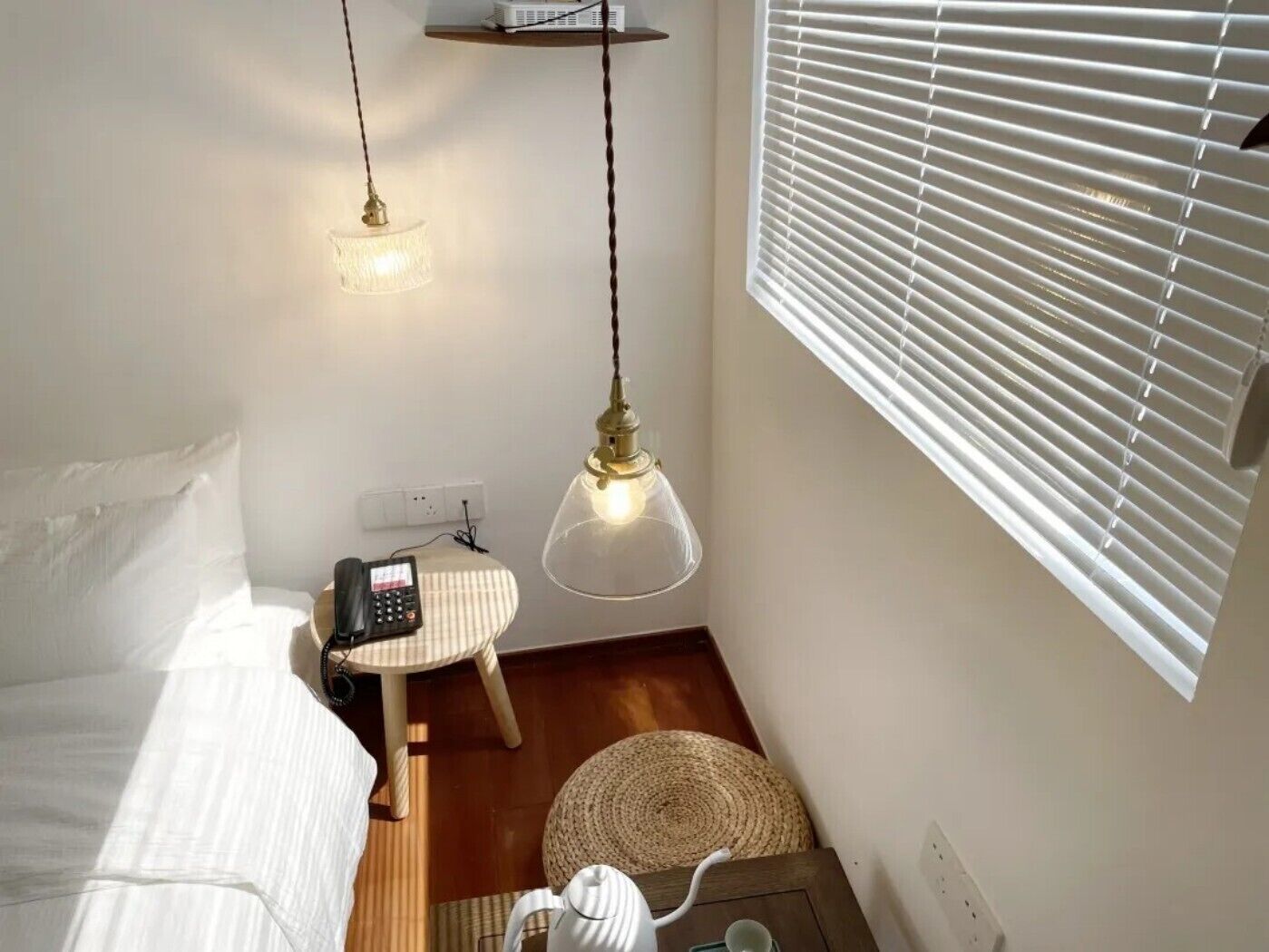 A small, cozy room with a bed, a desk with a telephone, a hanging light fixture, and a window with blinds.