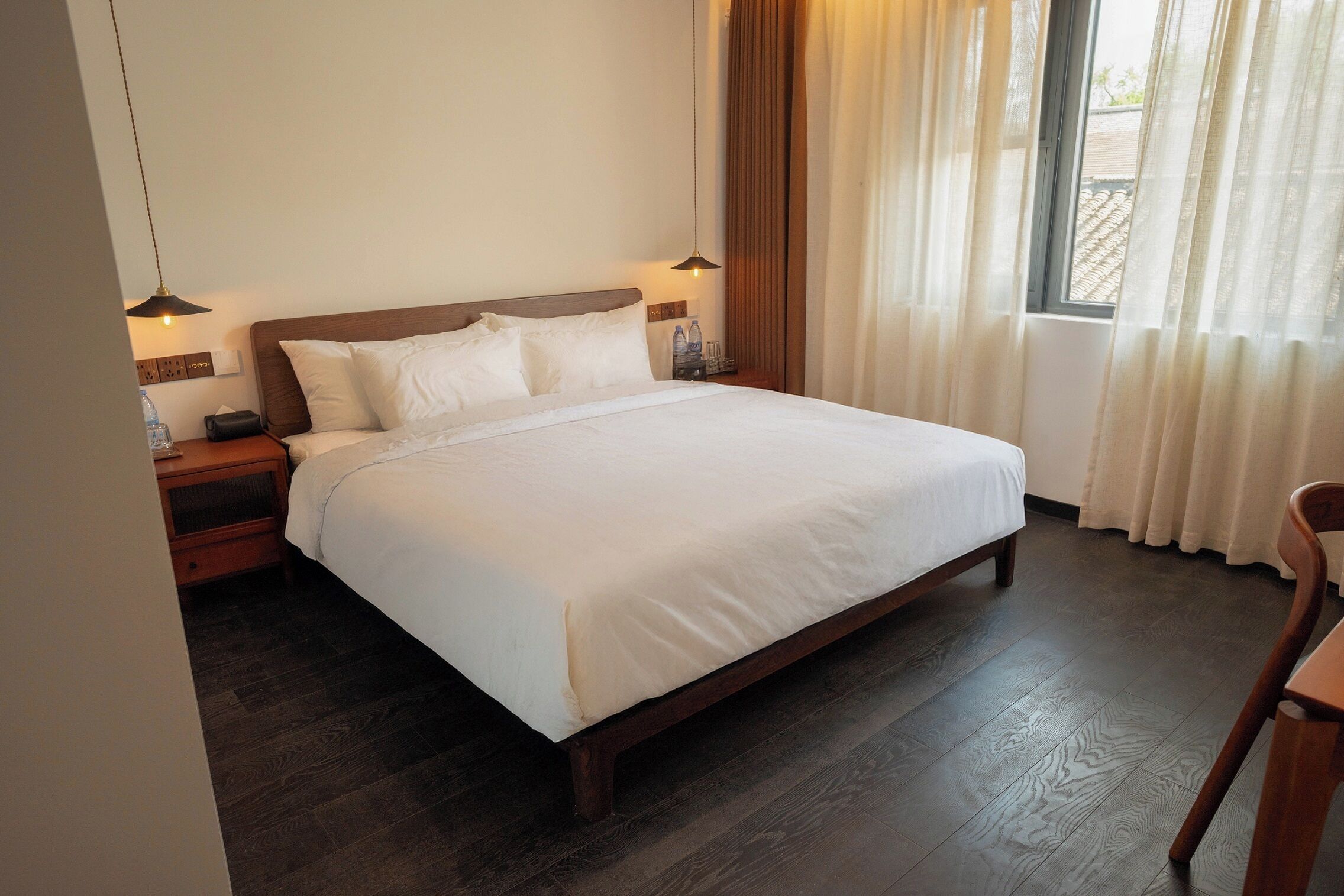 A neatly made bed with white linens, wooden headboard, two bedside tables with lamps, and a window with curtains.