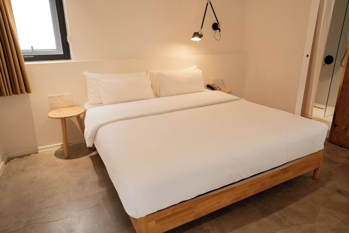 A neatly made bed with white linens, a bedside table, a lamp, and a window with curtains.