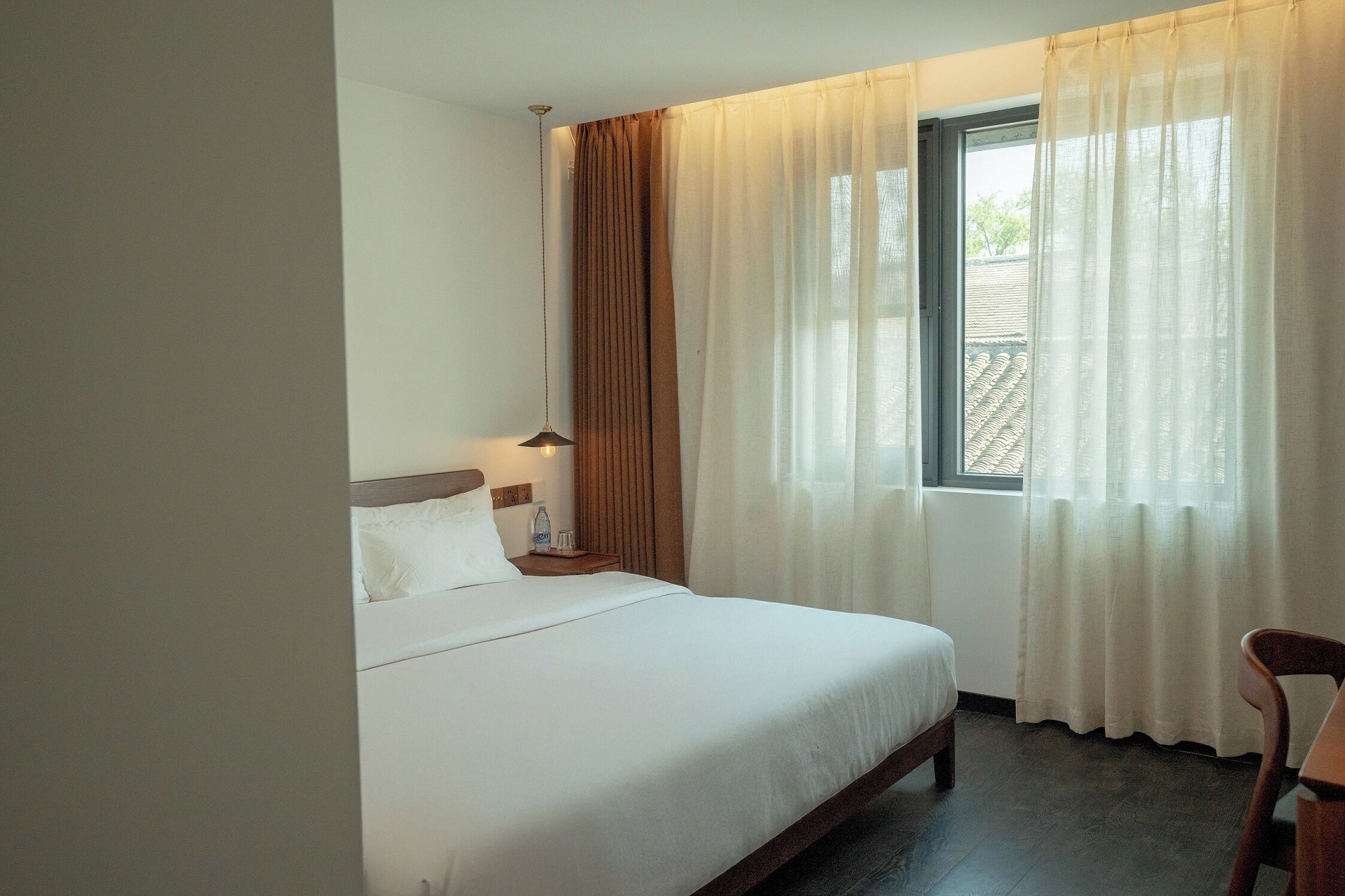 Una habitación de hotel con cama, mesita de noche, lámpara, ventana con cortinas y una silla.