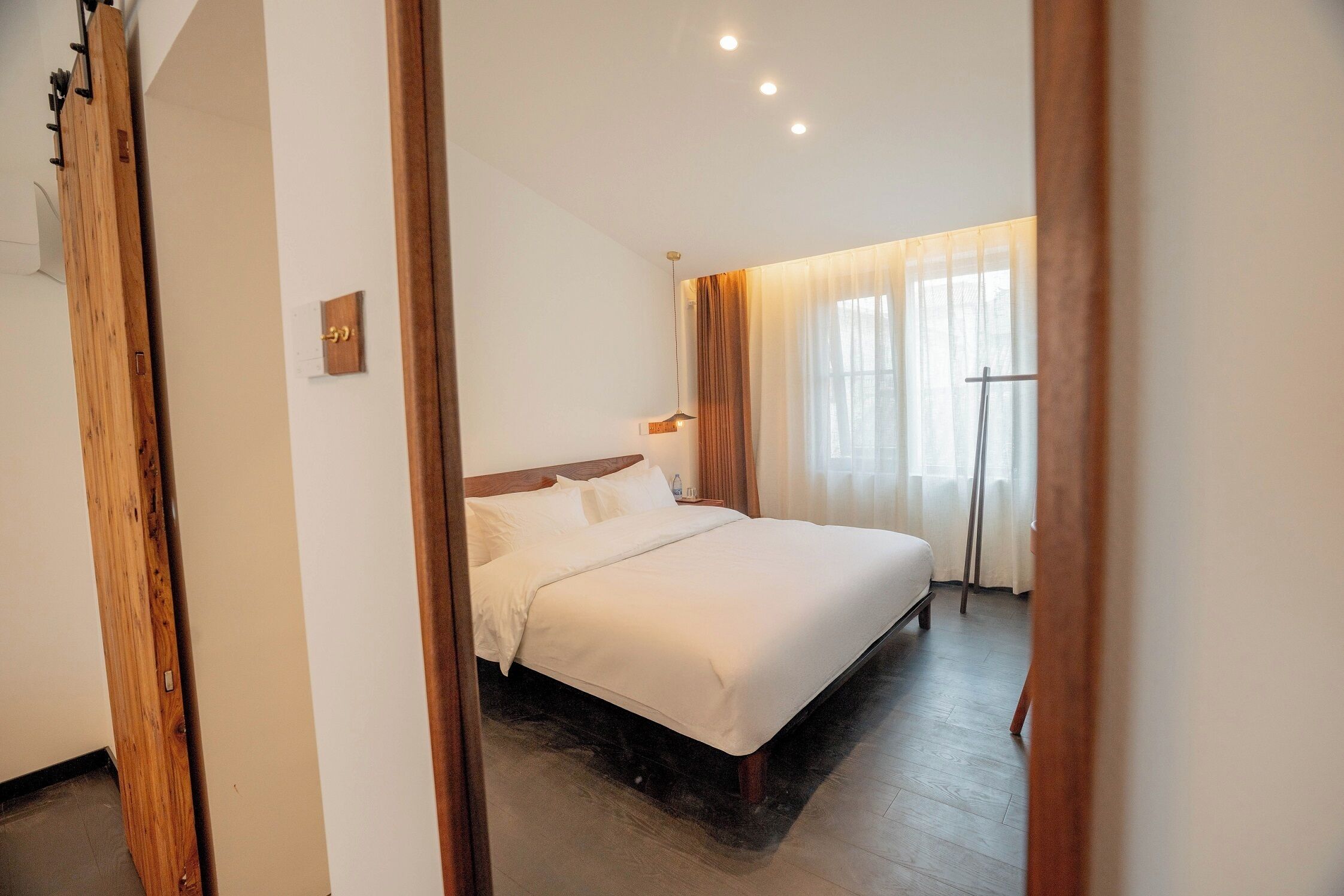 Habitación de hotel con cama, cabecero de madera, una mesita de noche, ventana con cortinas y una puerta.
