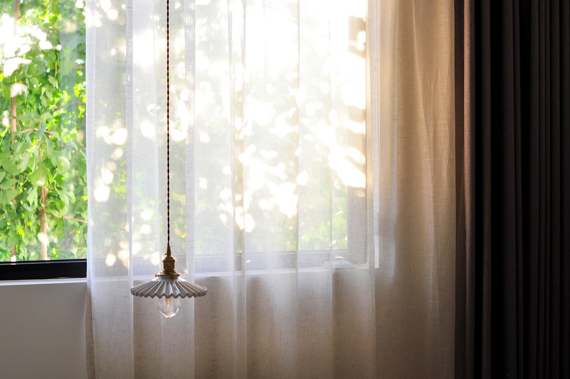 A room with a window covered by sheer curtains, a hanging light fixture, and a view of greenery outside.
