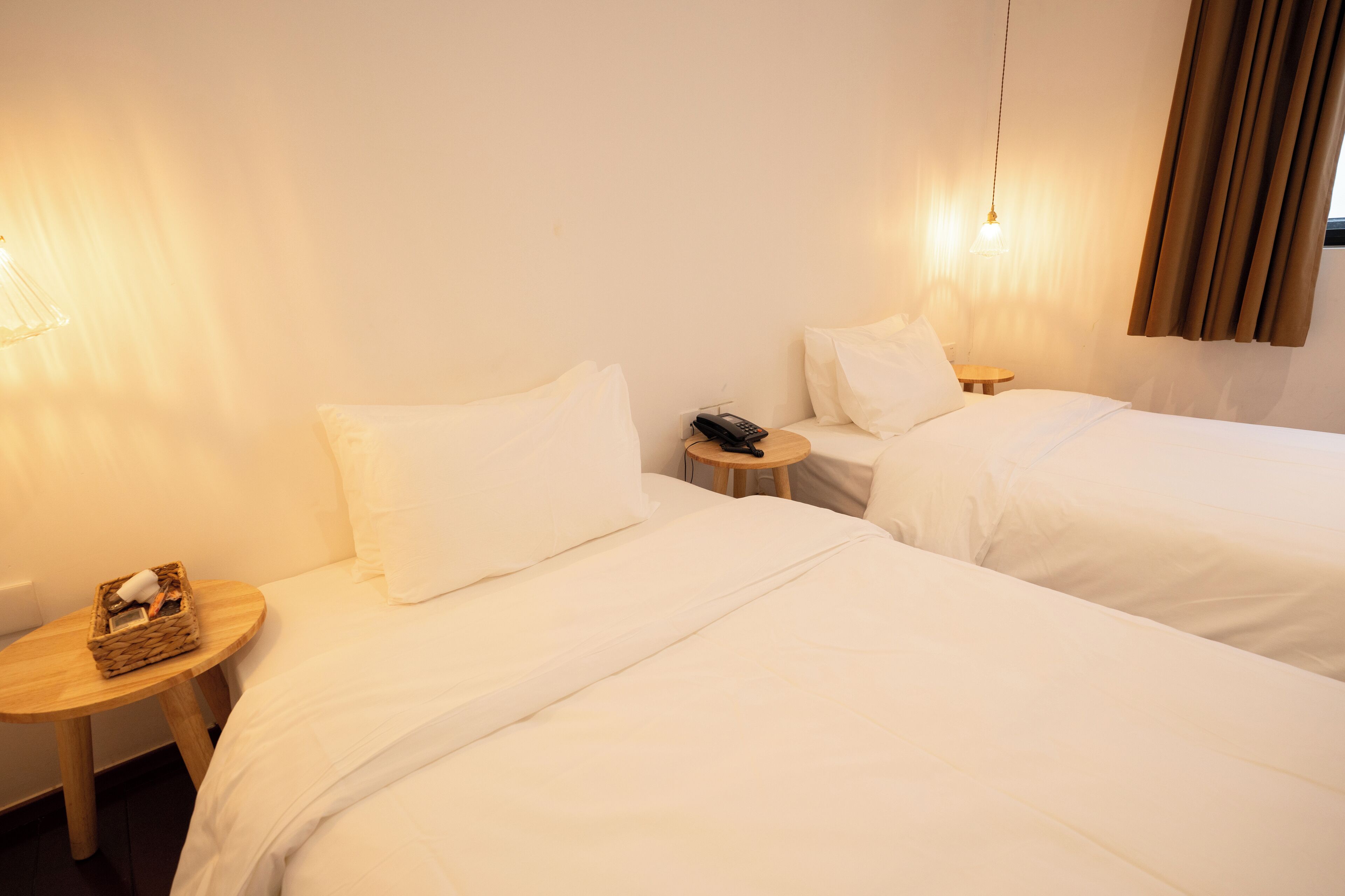 A hotel room with two beds, a bedside table with a lamp, and a small table with a basket.