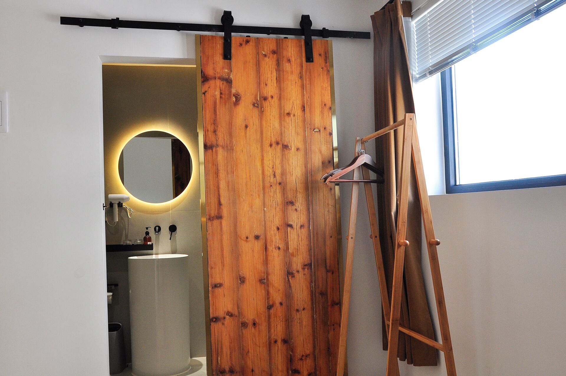 A room with a wooden sliding door, a mirror with a circular light, a wooden easel, and a window with blinds.