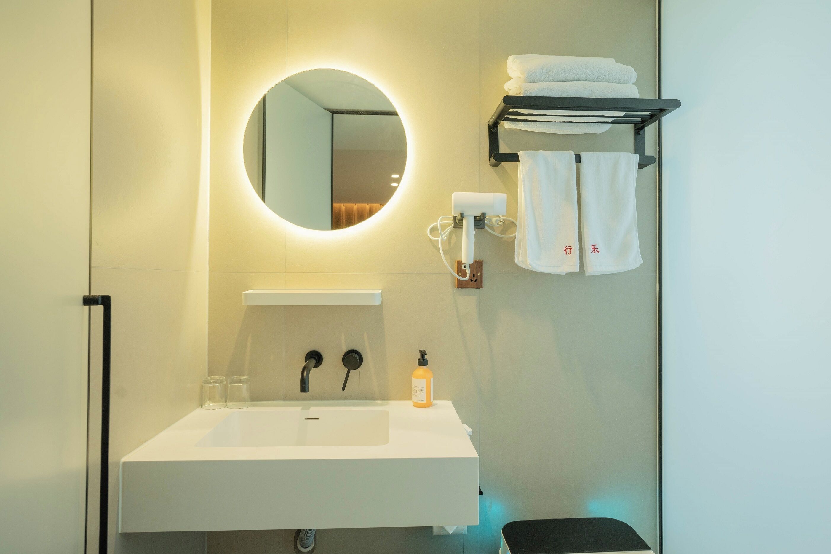 A hotel bathroom with a round mirror, a white sink, two faucets, a soap dispenser, and a towel rack with folded towels.