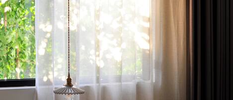 Ein Zimmer mit einem Fenster, das mit transparenten Vorhängen drapiert ist, einer Hängeleuchte und Blick auf Grünflächen.