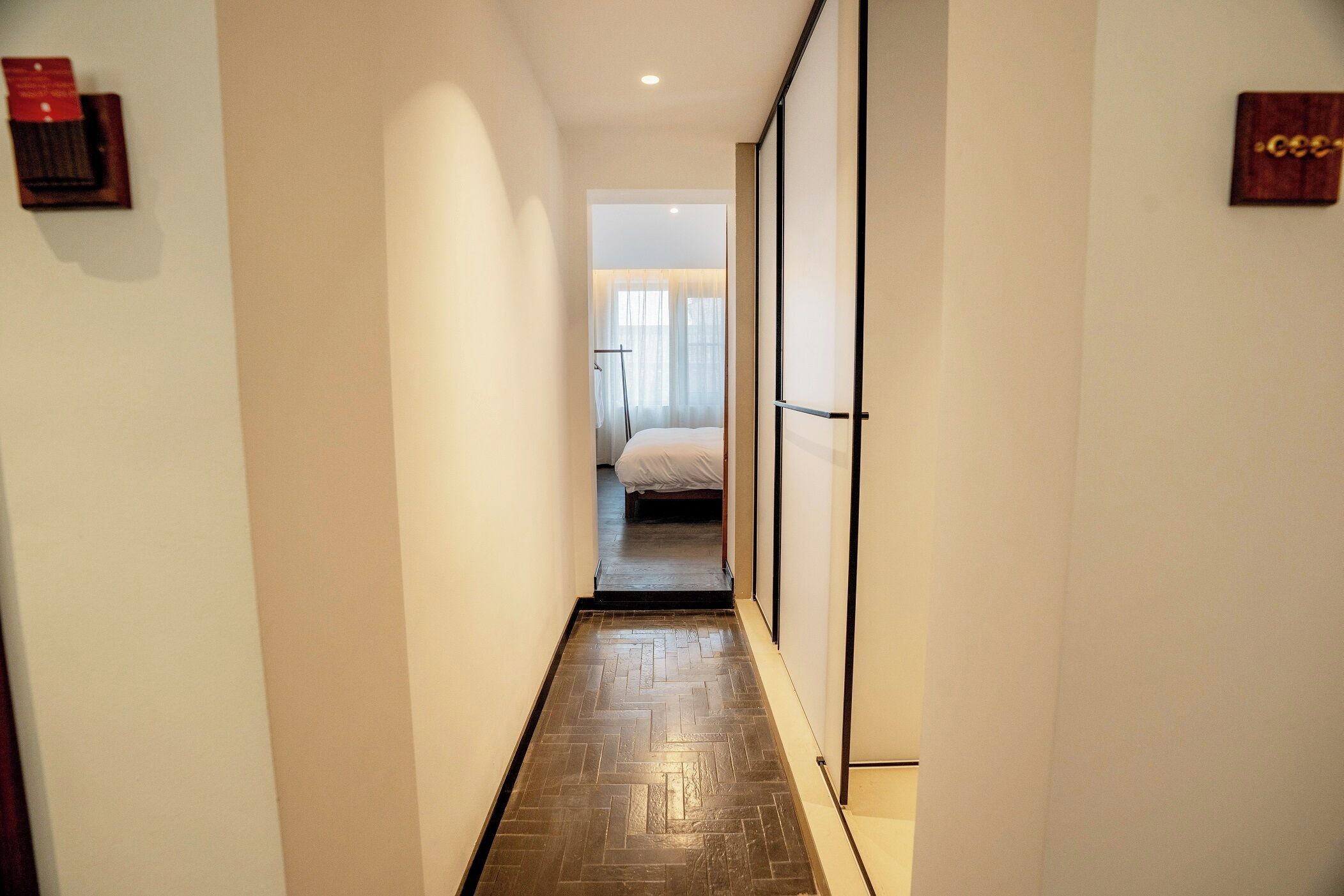 Un estrecho pasillo con suelo de madera que conduce a un dormitorio con cama y ventana.