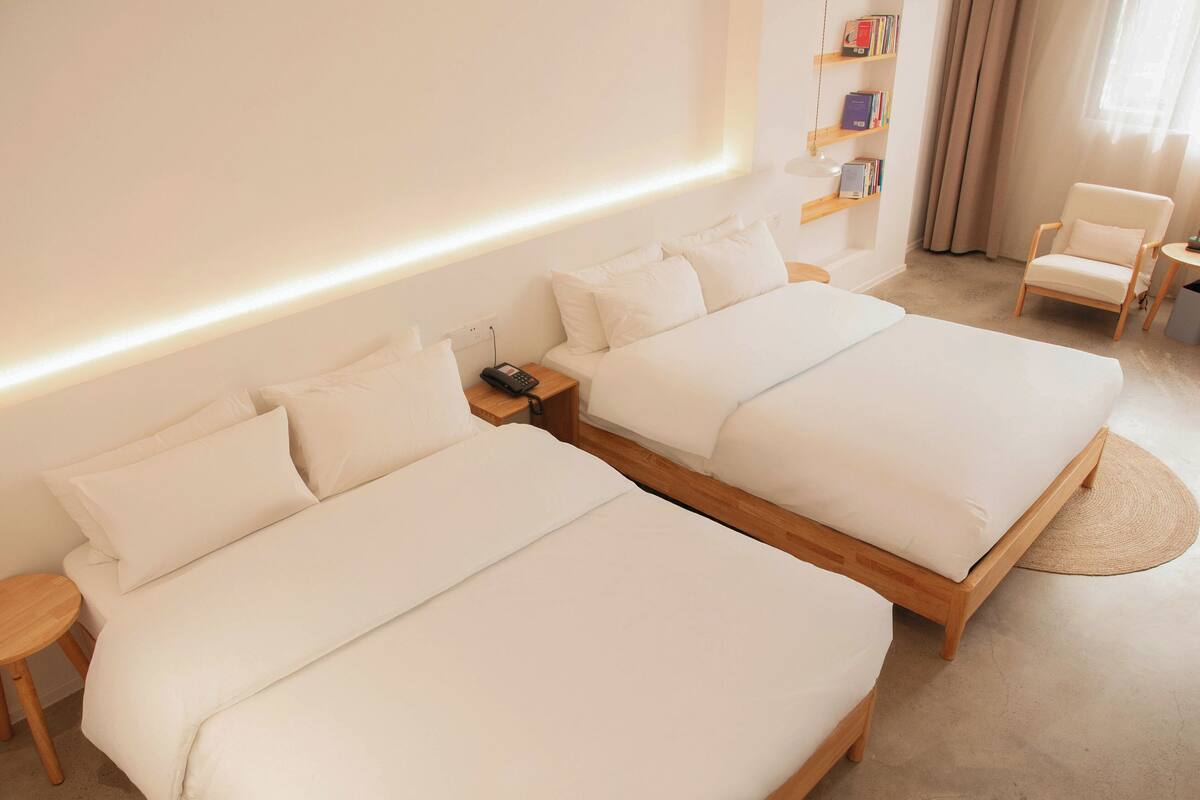 A hotel room with two beds, a bedside table with a phone, a small table with a lamp, a chair, and a bookshelf with books.