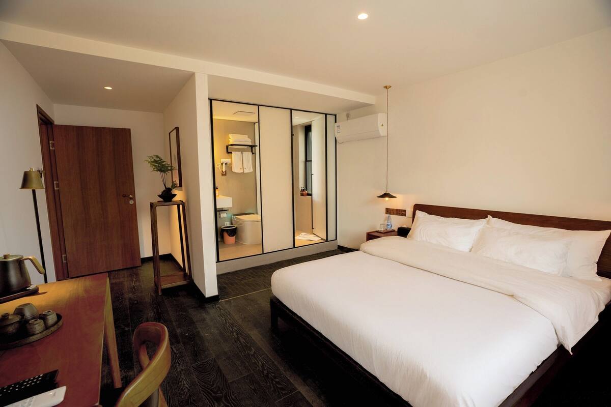 A hotel room with a bed, a desk with a chair, a small table with a lamp, and a mirror.